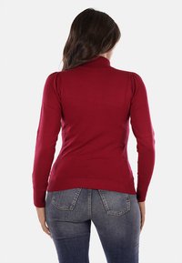 Maglione fitted rosso con collo alto, presenta polsini e fondo a coste e una texture liscia. Indossato con jeans in denim scuri.