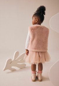 Gilet en fausse fourrure rose sur une chemise blanche à manches longues avec des petits motifs, jupe en tulle et chaussettes roses. Bottes de couleur claire avec une semelle épaisse.