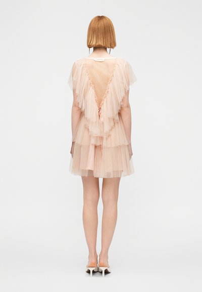 Γυναίκα με κοντά κόκκινα μαλλιά που φοράει στρωμένο φόρεμα από ροζ τούλι με βαθύ V στην πλάτη και λευκές γόβες, στέκεται μπροστά σε απλό λευκό φόντο.