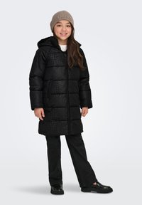 Smiling girl wearing a long black patterned puffer coat, black pants, black shoes, and a beige knit beanie on a plain light background.