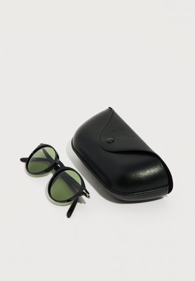 Μαύρα γυαλιά ηλίου με πράσινους φακούς δίπλα σε κλειστή μαύρη δερμάτινη θήκη με κουμπί κλίπς σε απλό λευκό φόντο.