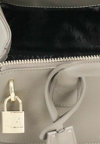 Borsa in pelle grigia con lucchetto dorato inciso "A X", cerniera parzialmente aperta che rivela una fodera interna nera con testo sbiadito.