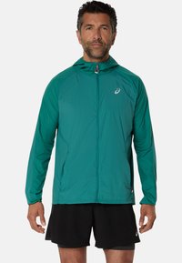 Veste à capuche légère couleur sarcelle avec fermeture éclair sur le devant, en tissu respirant et avec un logo discret sur la poitrine. Assortie à un short noir.