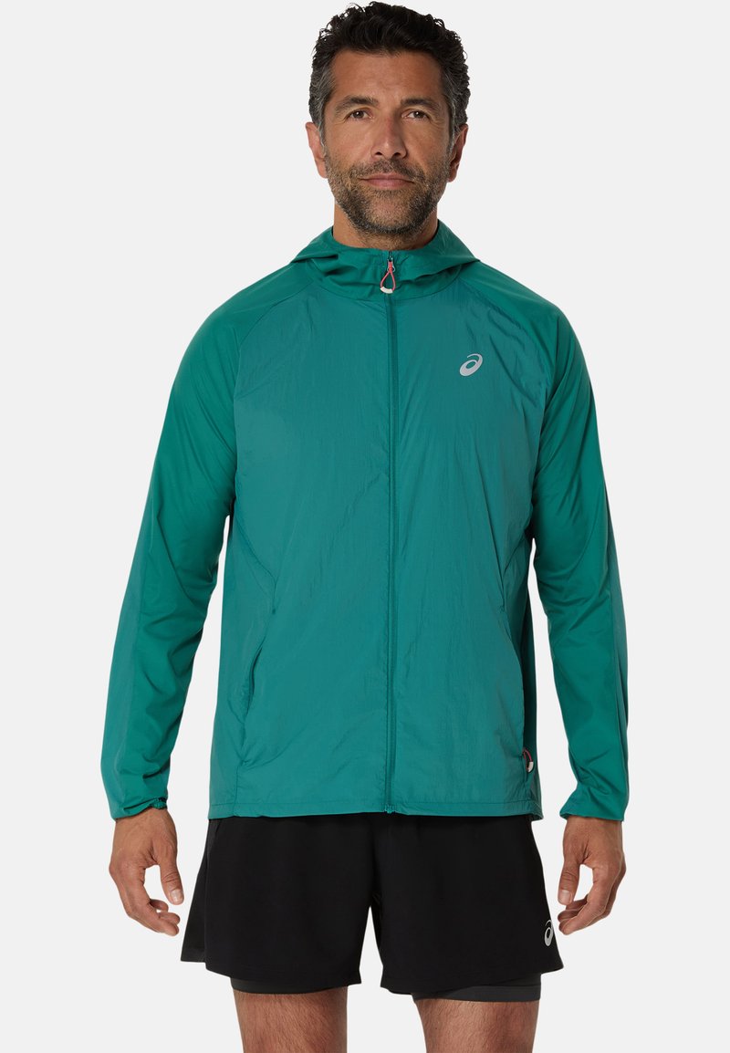 Veste à capuche légère couleur sarcelle avec fermeture éclair sur le devant, en tissu respirant et avec un logo discret sur la poitrine. Assortie à un short noir.