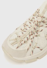 Ľahké tenisky s textúrovaným bielym sieťovinovým zvrškom, krémovými semišovými akcentmi, nastaviteľným systémom šnurovania a viazaniami s vzorom, ktoré majú červené a čierne vlákna.