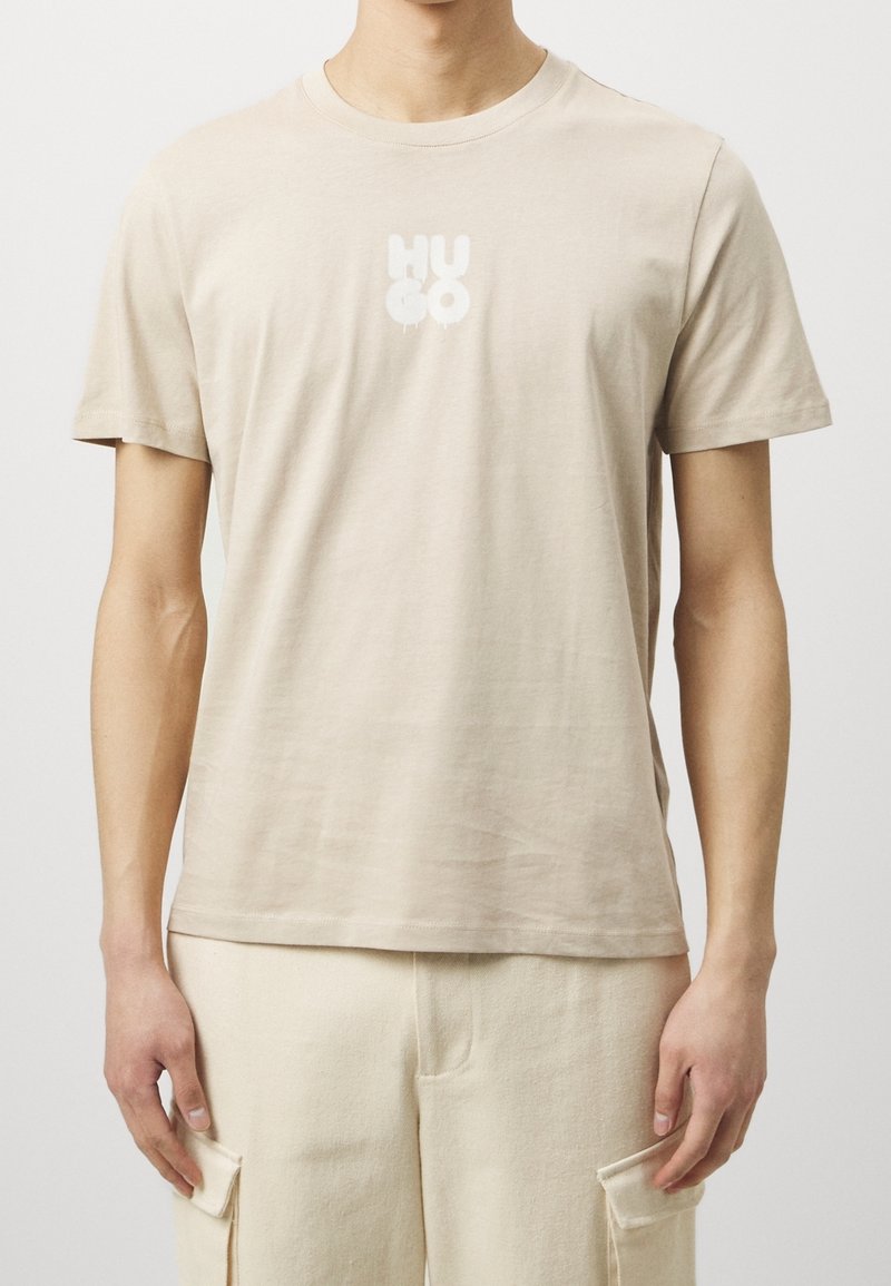 Beige cotton t-shirt with a crew neck and short sleeves. Features a white "HUGO" logo printed on the front. Smooth texture.