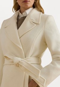 Garšs krēmu krāsas mētelis, izgatavots no mīksta vilnas, ar plašu apkakli, dubultā pogas josta un sasienamu jostu ap vidu.