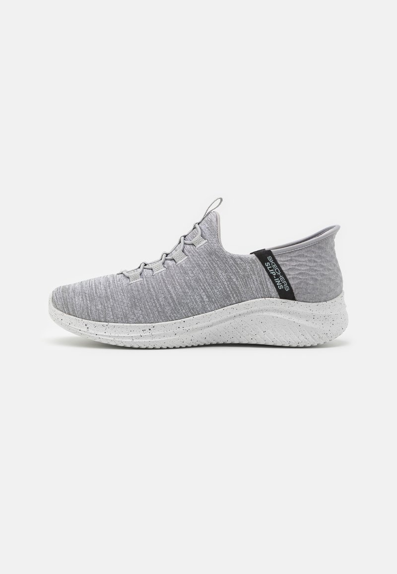 Chaussure de sport gris clair avec une tige texturée, sans lacets, et une semelle intermédiaire blanche avec des éclaboussures. Elle possède un onglet noir au talon pour un enfilage facile.
