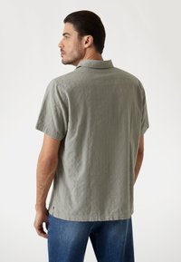 Camisa de manga corta en tela gris claro con patrón texturizado y collar con muesca, combinada con jeans azules, vista desde un ángulo trasero.