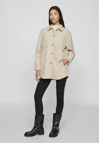 Giacca camicia in misto lana beige con due tasche sul petto e bottoni marroni, indossata sopra un maglione bianco. Abbinata a jeans skinny neri e stivali neri.