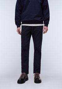 Mand iført sorte bukser, sorte sko med røde snørebånd, navyblå sweatshirt og lysegrå skjorte, stående mod en enkel lys baggrund.