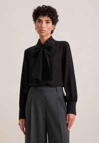 Schwarze Bluse mit einer großen Schleife, langen Ärmeln und geknöpften Manschetten. Kombiniert mit grauen, maßgeschneiderten Hosen aus strukturiertem Stoff.