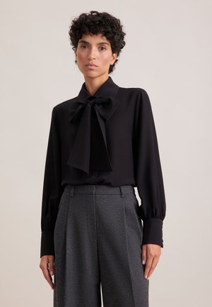 Blusa nera con un grande fiocco, maniche lunghe e polsi abbottonati. Abbinata a pantaloni grigi su misura con tessuto texturizzato.