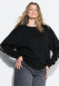 Czarny, oversize sweter dziany z prążkowanym dekoltem i mankietami, z dekoracyjnymi srebrnymi akcentami na rękawach. Noszony z szarymi dżinsami.