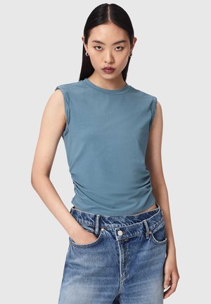 Młoda kobieta o długich ciemnych włosach, ubrana w niebieski bezrękawnik i wyblakłe niebieskie dżinsy z wysokim stanem, z jedną ręką w kieszeni.