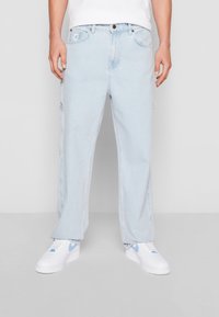 Jeans de denim azul claro con un corte relajado, que presentan un cierre de botón, bolsillos frontales y un pequeño parche de marca en la cintura.
