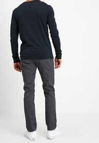 Zwart shirt met lange mouwen en ronde hals, gecombineerd met grijze broeken en witte sneakers. Het shirt heeft grafische ontwerpen op de mouwen.