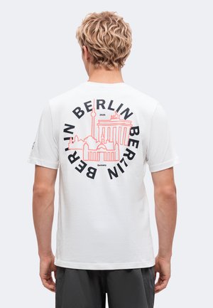 Homme aux cheveux bouclés portant un t-shirt blanc avec une silhouette rose de Berlin et un texte noir "BERLIN" en cercle dans le dos.