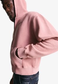 Lyserød zippered hoodie lavet af blødt, tekstureret stof. Har en frontlomme og ribbede manchetter, båret med mørke jeans.