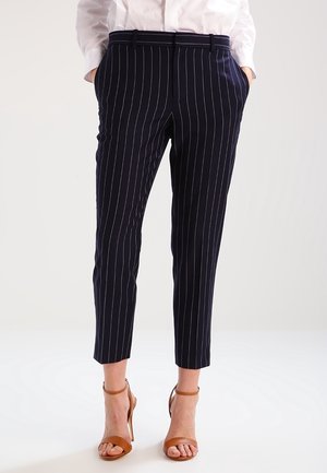 Femme portant un pantalon court bleu marine à fines rayures avec poches et des sandales à talons hauts beige, debout devant un fond uni.