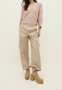 Frau trägt einen hellrosa strukturierten Strickpullover, beige Cargo-Hosen mit elastischen Bündchen und beige Plateausandalen, steht vor einem schlichten Hintergrund.