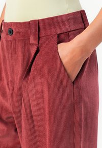 Mano nella tasca di pantaloni bordeaux a vita alta, plissettati e con texture a coste, con bottone nero e passanti per cintura, indossati con una maglietta beige.