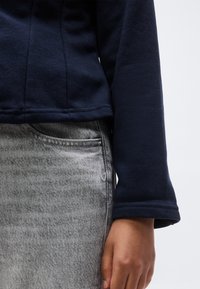 Άτομο που φοράει μπλε σκούρη μπλούζα με μακριά μανίκια και ανοιχτό γκρι τζιν φούστα, με το χέρι του να ακουμπά στο πλάι.