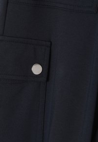 Gros plan d'une poche en tissu sombre avec rabat sécurisé par un bouton-pression rond argenté sur un vêtement.