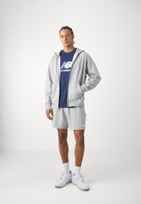 Grå zip-hoodie over en marineblå T-shirt, parret med grå shorts og hvide sneakers. Simpelt design med logo detaljer på tøjet.