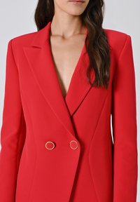 Donna che indossa un blazer rosso aderente a doppio petto con bottoni circolari dorati e lunghi capelli scuri e ondulati che le scendono sulla spalla.