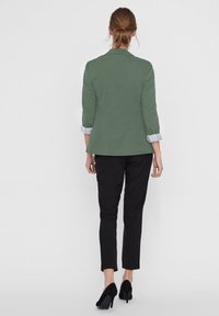 Zelený ušitý sáčko s ohrnutými světlomodrými rukávy, v kombinaci s černými úzkými kalhotami a černými vysokými podpatky, pohled zezadu.