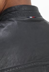 Achteraanzicht van een persoon die een zwarte leren jas draagt met een gestructureerde kraag en een klein rood, wit en blauw stoffen label bij de hals.