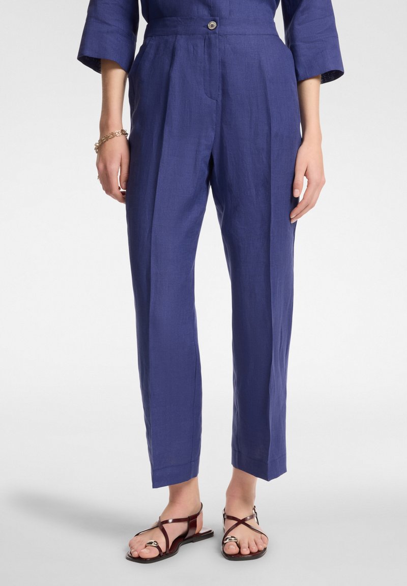 Pantaloni in lino blu navy con vestibilità rilassata, dotati di chiusura anteriore con bottone, tasche laterali e dettagli plissettati che scorrono sulla parte anteriore.