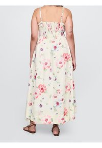 Femme portant une robe maxi fleurie crème avec de fines bretelles et des sandales plates marron, vue de dos sur un fond uni.