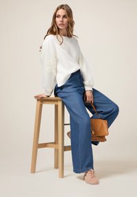 Sweatshirt blanc avec des accents en dentelle, pantalon en denim bleu à jambes larges, baskets roses pâles et un sac à main marron, assis sur un tabouret en bois.