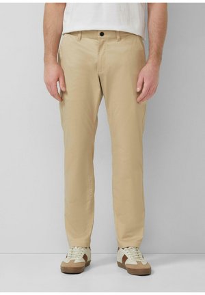 Man draagt beige broek met rechte pijpen, witte sneakers met bruine accenten en een wit overhemd, staand tegen een eenvoudige lichte achtergrond.