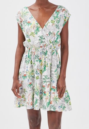 Femme portant une robe blanche sans manches à imprimé floral avec décolleté en V et taille cintrée, posant devant un fond clair uni.