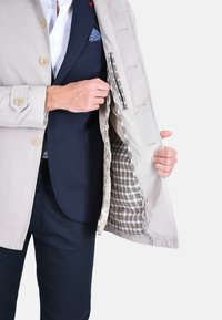 Cappotto beige chiaro con bottoni, con fodera a quadri. Tasca interna visibile, abbinato a un blazer blu navy. Tessuto dalla texture liscia.