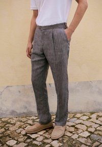 Pantalons gris à rayures avec une coupe ajustée, présentant une taille plate, des poches latérales et un éclat subtil. Portés avec des chaussures beiges à enfiler.
