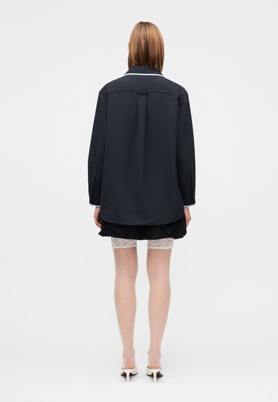 Pārmēra lieluma tumši zila krekla ar apkakli, garām piedurknēm un pogu aprocēm, valkāts pāri melnai svārkiem ar ziedu akcentiem un mežģīņu apdari.