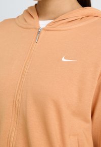 Persikofärgad zip-hoody med mjuk textur, med en vit Nike-logga på bröstet och en klassisk dragsko i huvan.