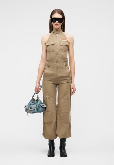 Femme en combinaison sans manches couleur sable tenant un sac à main bleu, portant de grandes lunettes de soleil noires et des bottes noires, debout devant un fond uni.