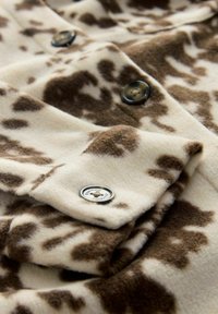 Fleece jacket in cream and brown animal print, featuring dark buttons and a silver logo button. Soft texture with a warm appearance.