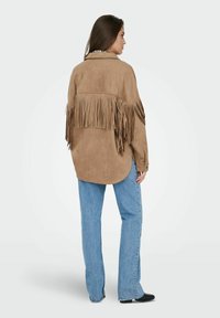 Brun ruskindsjakke med frynser på bagsiden, kombineret med lyseblå straight-leg jeans og sorte sko. Enkel krave og knaplukning ved manchetterne.