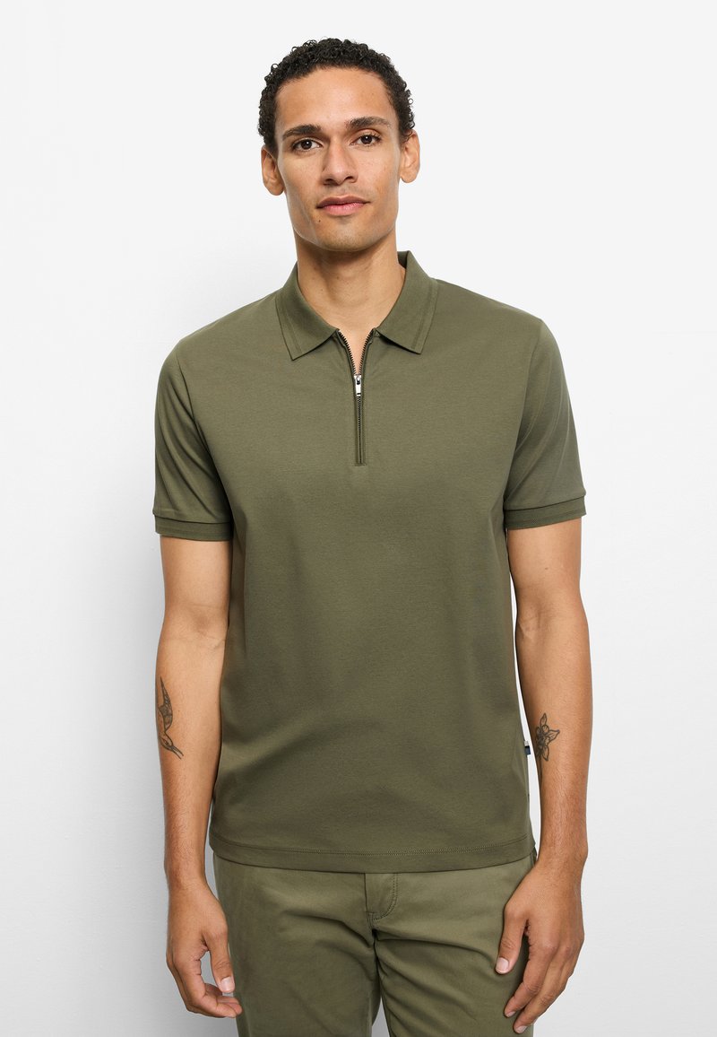 Mann steht und trägt ein olivgrünes kurzärmeliges Poloshirt mit Frontreißverschluss und passende Hose vor weißem Hintergrund.