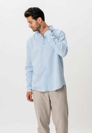 Mężczyzna w jasnoniebieskiej koszuli na guziki i beżowych spodniach delikatnie dotykający szyi, patrzący w dół na jednolitym białym tle.