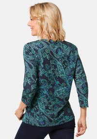 Langarmbluse aus dunklem Stoff mit einem Paisleymuster in Grüntönen und Blau. Mit Raffung an den Schultern und einem runden Ausschnitt.