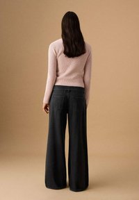 Femeie cu păr lung și închis la culoare, purtând un pulover roz deschis cu model tip nervuri și blugi negri cu picioare largi, stând cu fața spre un fundal bej, cu spatele întors.