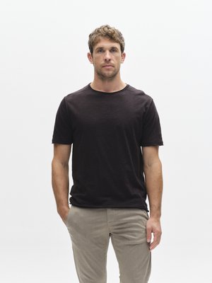 Camiseta de color marrón oscuro con cuello redondo, hecha de tela suave. El modelo lleva pantalones beige claro. Diseño sencillo, textura lisa y mangas cortas.