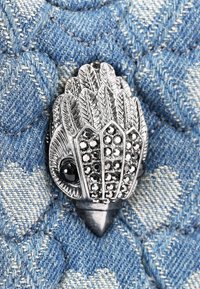 Broche en argent en forme d'oiseau avec des motifs de feuilles détaillés, ornée de pierres noires, posée sur un fond en denim bleu texturé.